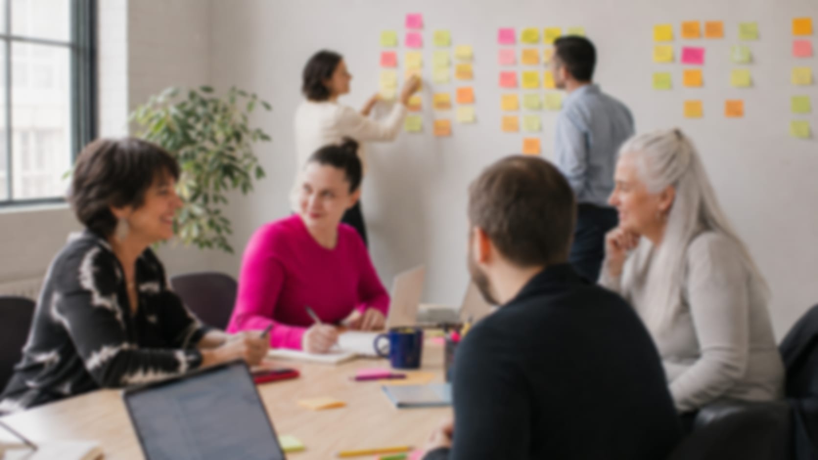 Diverses Team im Workshop: eine Person heftet Ideen an eine Wand voller bunter Haftnotizen, Kolleginnen und Kollegen arbeiten mit Laptops am Tisch zusammen