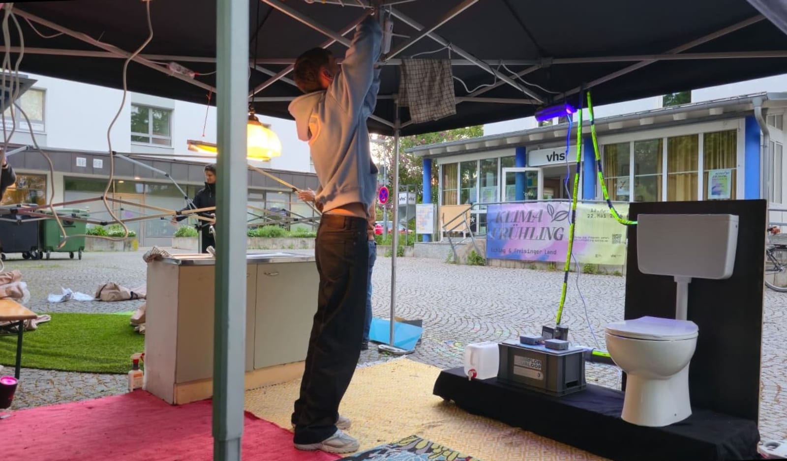 Aufbau bei der Open-Air-Veranstaltung Klimafrühling: Menschen richten unter einem Pavillon eine Ausstellung ein, im Hintergrund ein Banner der Veranstaltung